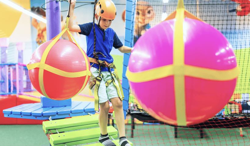 A young boy walking across a bridge at an obstacle course with a harness and a helmet